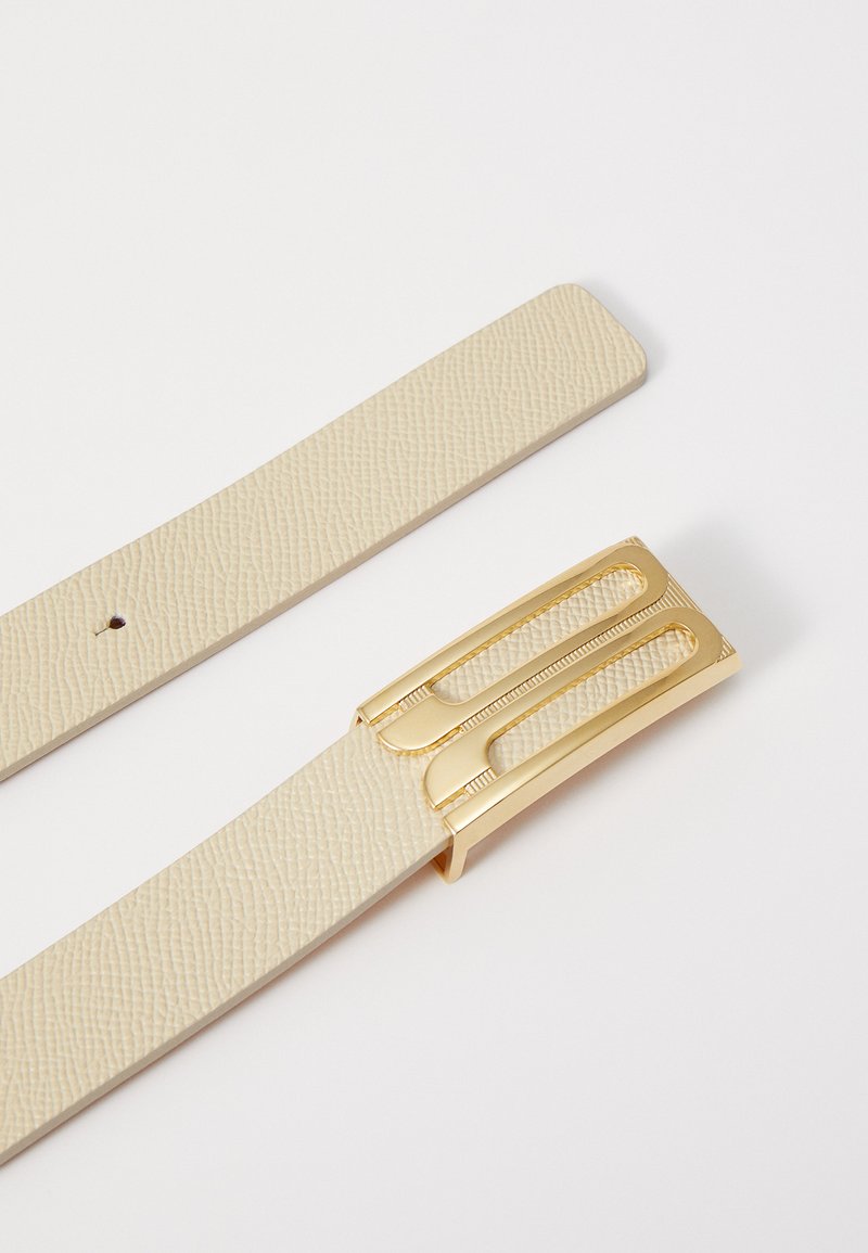 Cinturón de cuero beige con acabado texturizado y una hebilla de metal dorado que presenta un diseño recortado y líneas verticales.