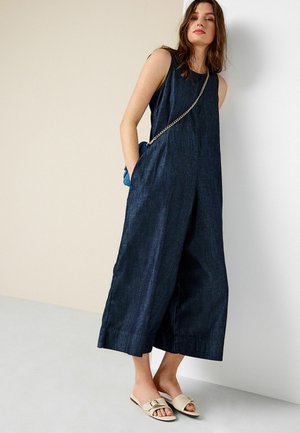 Kobieta stojąca przy ścianie, ubrana w ciemny, bezrękawowy kombinezon z denimu, beżowe klapki i małą niebieską torebkę z złotym łańcuszkiem.