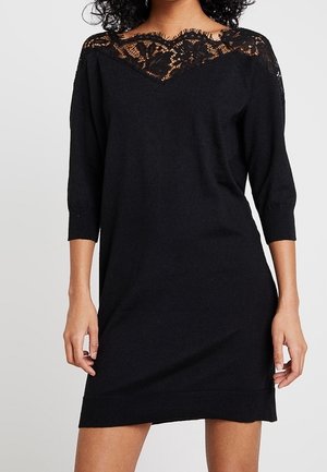 Femme portant une robe noire longueur genou avec des manches trois-quarts et un détail en dentelle au niveau du col et des épaules.