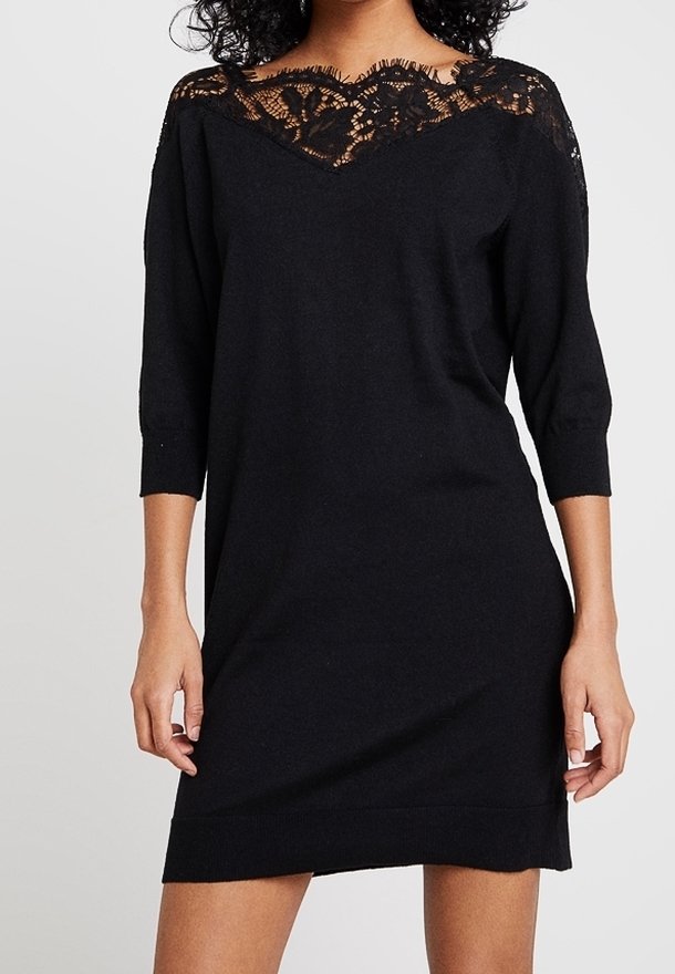 Femme portant une robe noire longueur genou avec des manches trois-quarts et un détail en dentelle au niveau du col et des épaules.