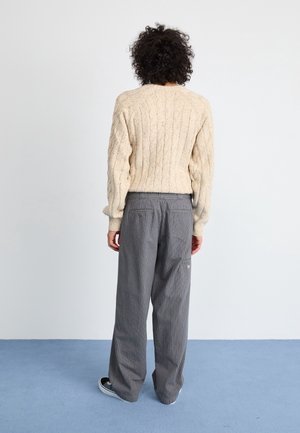 Person mit lockigem Haar, die einen beigen, kabelgestrickten Pullover, graue gestreifte Hosen und schwarze Turnschuhe trägt und einer weißen Wand gegenübersteht.