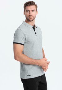 Polo de color gris claro con cuello y puños negros, que presenta una textura suave y un discreto bordado del logo en el dobladillo.