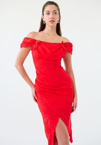 Robe rouge décolletée avec des nœuds aux manches, au design ajusté et à tissu texturé. Présente une fente haute sur un côté.