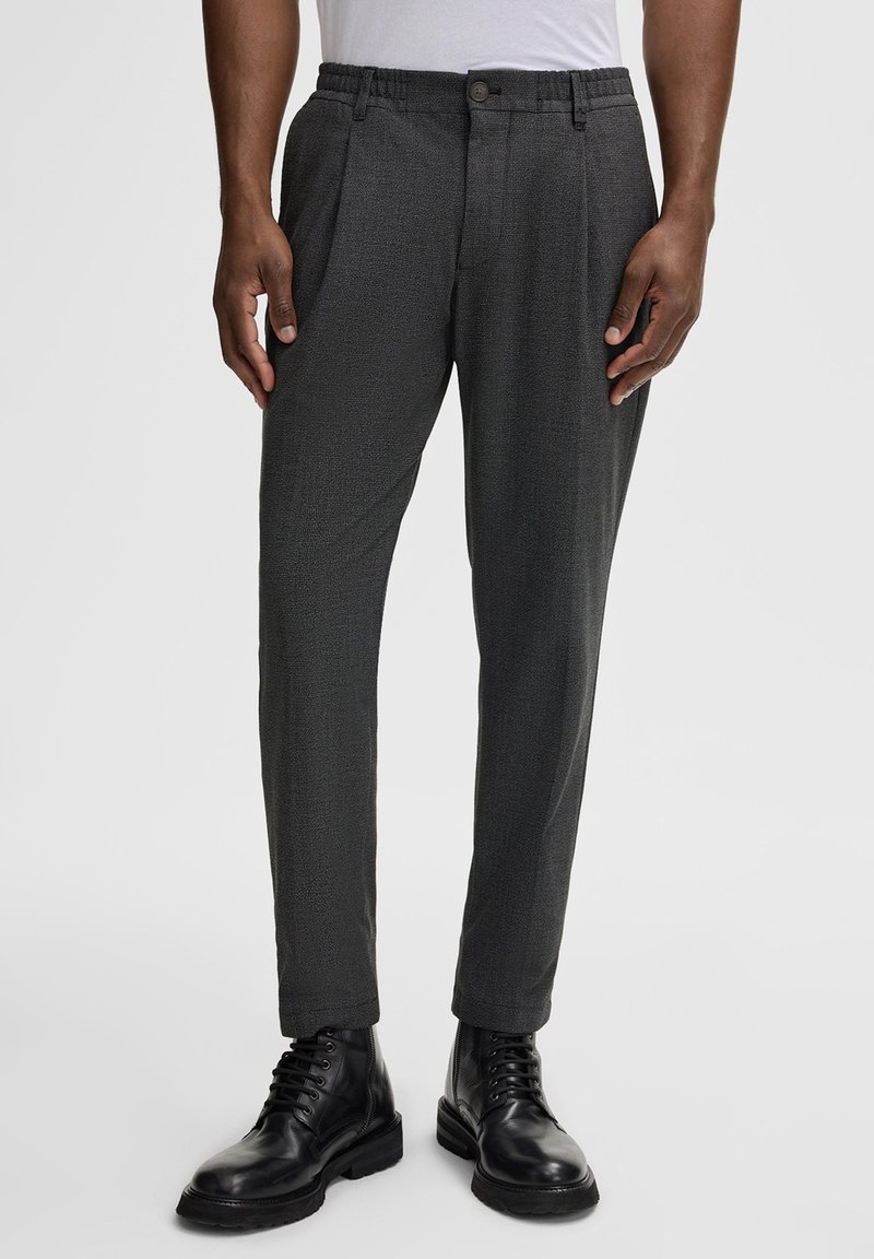 Pantalons plissés gris foncé en tissu texturé avec une taille élastique, dotés d'une fermeture à bouton sur le devant et de jambes cintrées. Portés avec des bottes noires.