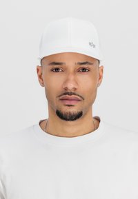 Cască albă din țesătură netedă, cu o vizieră curbă și logo argintiu. Purtați cu un tricou alb simplu, cu guler rotund. Design curat, minimalist.