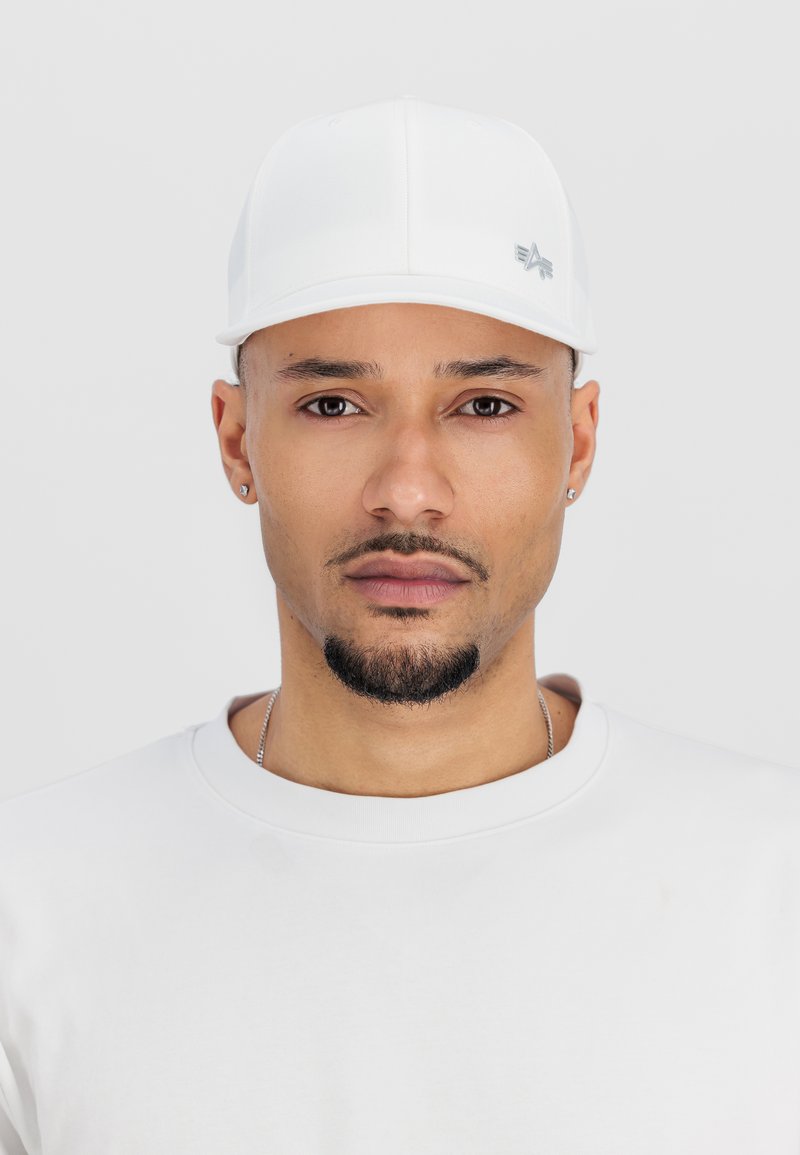 Cască albă din țesătură netedă, cu o vizieră curbă și logo argintiu. Purtați cu un tricou alb simplu, cu guler rotund. Design curat, minimalist.