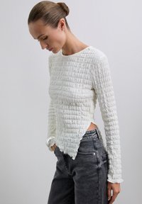 Kobieta z włosami upiętymi w kok, ubrana w fakturowany biały top z długim rękawem i czarne jeansy z wysokim stanem, stojąca z rękami w kieszeniach na jednolitym tle.