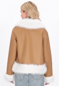 Blonde Frau mit einer beigefarbenen, gefälschten Lederjacke, die einen weißen Kunstfellkragen, -manschetten und -saum hat, kombiniert mit hellblauen Jeans, Rückansicht.