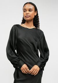 Melna satīna blūze ar garām piedurknēm un laivu veida kakla izgriezumu, ar puffed piedurknēm un piegulošām aprocēm. Iekļauj smalku mirdzumu un dekoratīvas detaļas uz piedurknēm.