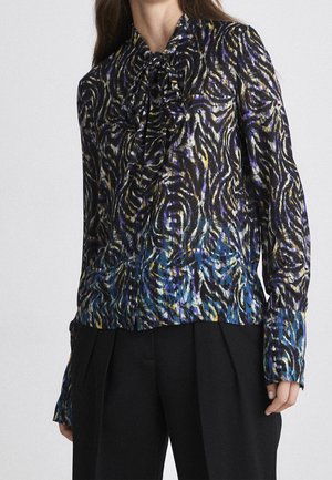 Femme portant un chemisier à manches longues avec un imprimé animal abstrait multicolore et un col lavallière, associé à un pantalon noir plissé.