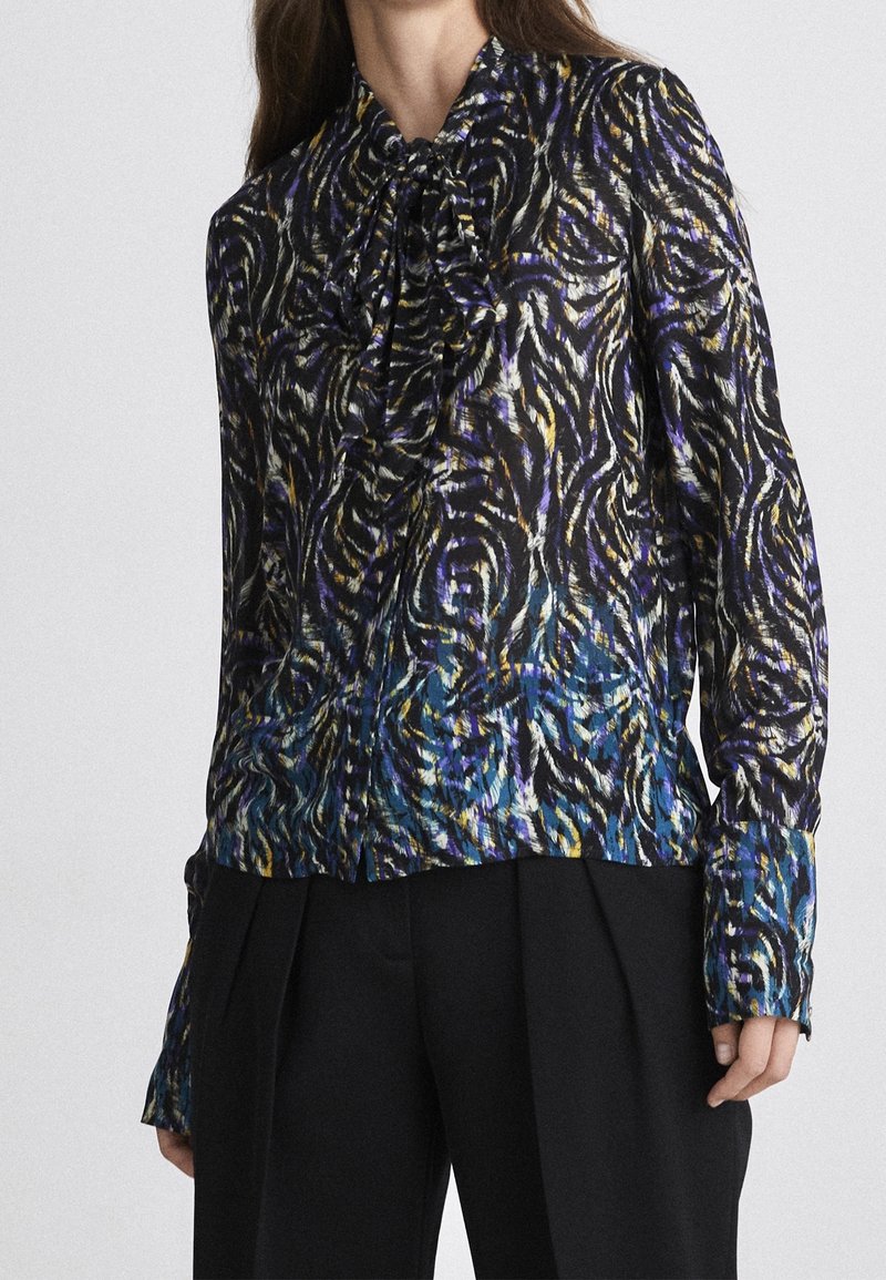Femme portant un chemisier à manches longues avec un imprimé animal abstrait multicolore et un col lavallière, associé à un pantalon noir plissé.