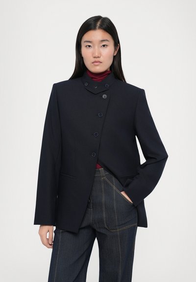 Marineblazer met een hoge kraag, zes knopen aan de voorkant en twee zijzakken. Gedragen over een bordeauxkleurige top en gecombineerd met denim broek met wijde pijpen.