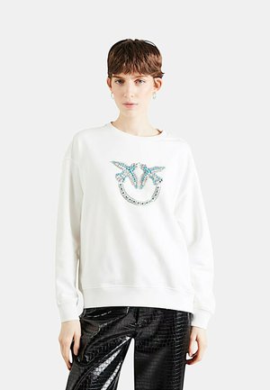 Felpa bianca con logo decorativo in cristallo blu e argento sul davanti. Il modello indossa pantaloni neri, lucidi e texturizzati.