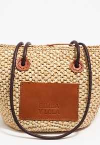 Bolso de paja tejido con un acento de cuero marrón y parche de logo. Presenta una forma redondeada, asas de cuerda marrón oscuro y una textura natural.