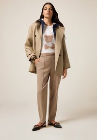 Beige trenchcoat met marineblauwe voering, witte trui met een tan beerafbeelding, tan op maat gemaakte broek, en zwarte flats met een geweven detail.