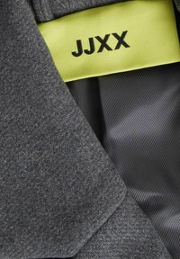 Sivi materijal s mekom teksturom, sa svijetlo žutom oznakom s natpisom "JJXX" i glatkom crnom podlogom ispod.