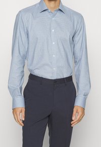 Uomo che indossa una camicia da uomo a maniche lunghe di colore azzurro chiaro con motivo, infilata in pantaloni eleganti blu scuro e con una cintura nera.