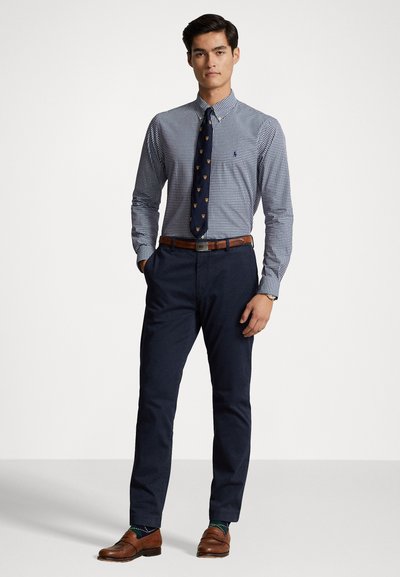 Chemise à boutons en motif vichy bleu et blanc, cravate marine avec de petites accents dorés, ceinture marron, pantalon foncé et mocassins marron.