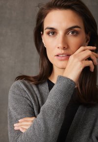 Cardigan en tricot gris avec des manches larges, porté sur un haut noir. Le mannequin a de longs cheveux bruns et un maquillage subtil, avec une main reposant sur son visage.