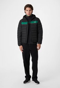 Sort pufferjakke med vandrette grønne og røde striber, lynlåslukning og hætte, kombineret med mørke bukser og sorte sneakers. Glat tekstur.