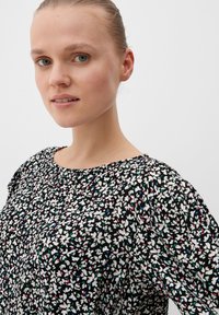 Schwarzbluse mit floralem Muster, die kleine weiße Blumen mit bunten Akzenten zeigt. Lockere Passform, runder Ausschnitt und strukturierter Stoff.