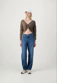 Top cropped de malha castanho com um design torcido, combinado com jeans azul desfiado e ténis brancos; modelo a usar um boné branco.