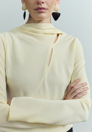 Mujer con top amarillo pálido de escote asimétrico y recortes, brazos cruzados y pendientes colgantes geométricos bicolor.
