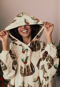 Hættet tæppe med cremefarvet stof og mønstre af julepudding og kristtornblade. Elastiske manchetter og blød tekstur giver varme.