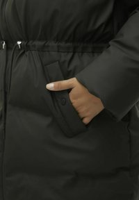 Veste doudoune noire avec taille cintrée, poche latérale à fermeture à pression et texture lisse et mate. Main reposant à l'intérieur de la poche.