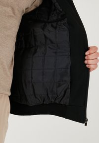 Sort jakke med en kviltet sort inderside. Har en ribbet kant, sidelommer med lynlås og en glat stofstruktur.