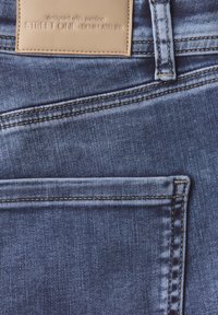 Jean en denim avec un dégradé de bleu clair à foncé, présentant des coutures visibles, une étiquette en cuir beige et une poche avant.