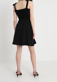 Černé áčkové šaty se smockovaným živůtkem, nastavitelnými ramínky s mašlemi a hladkou texturou látky. Nošeny s černými sandály na podpatku.
