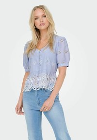 Blouse bleu clair à manches bouffantes, avec des motifs floraux brodés, un col en v et un ourlet en dentelle blanche froncée, associée à un jean en denim clair.