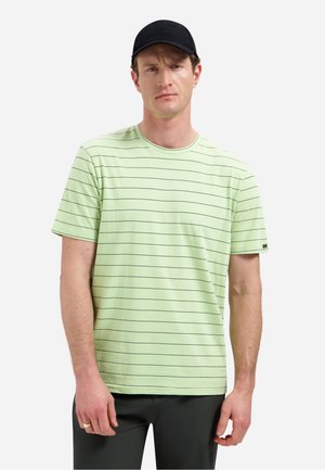 Man draagt een lichtgroen T-shirt met korte mouwen en dunne horizontale strepen, zwarte broek en een zwarte pet, staand tegen een effen achtergrond.