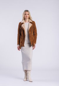Veste en daim marron avec un col en fourrure, assortie à une robe moulante gris clair et des bottes glossy beiges jusqu'aux genoux.