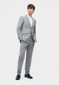 Νέος άνδρας που φοράει ανοιχτό γκρι κοστούμι με άσπρο πουκάμισο και μαύρα παπούτσια, στέκεται με τα χέρια στις τσέπες μπροστά σε λευκό φόντο.