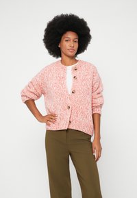Mujer con abundante cabello rizado que lleva un cárdigan de punto rosa y blanco con botones de madera sobre una blusa blanca y pantalones verde oliva, de pie con la mano en la cadera.