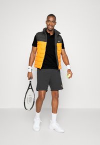 Pohodlná vesta v šedé a oranžové barvě, černé polo tričko, šedé kraťasy, bílé ponožky a tenisky; drží tenisovou raketu a míček.