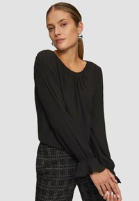 Schwarze Bluse mit langen Ärmeln und gefaltetem Design, mit gerafften Manschetten und runder Ausschnitt, kombiniert mit strukturierten schwarz-weißen karierten Hosen.