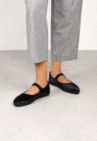 Zapatos Mary Jane negros, hechos de material suave, con suela plana, punta redonda y una correa a través del empeine. Acabado en tela texturizada.