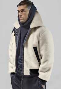 Jachetă din fleece de culoare crem, cu o textură de tip shearling, accente negre și fermoar. Pur tată pe un hanorac închis la culoare, cu un guler înalt.