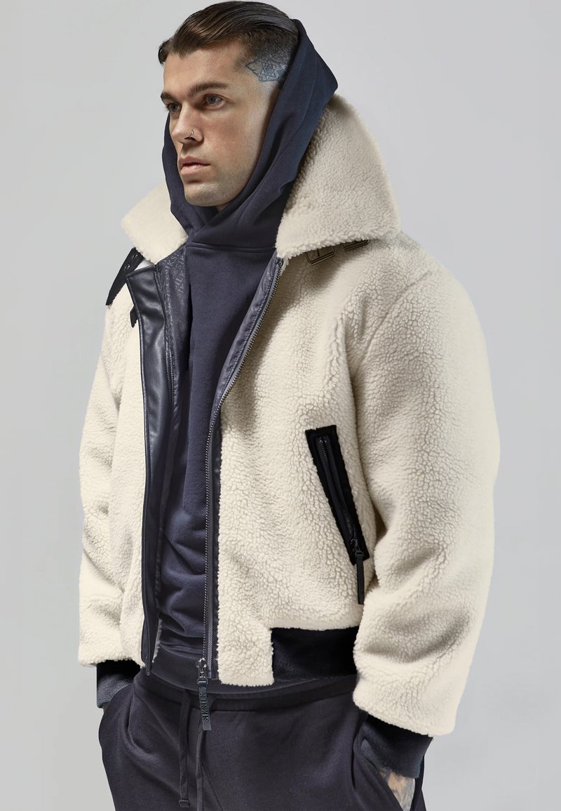 Jachetă din fleece de culoare crem, cu o textură de tip shearling, accente negre și fermoar. Pur tată pe un hanorac închis la culoare, cu un guler înalt.