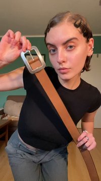 Brauner Leder-Gürtel mit einer silbernen rechteckigen Schnalle. Der Gürtel ist breit und glatt und wird von einer Person getragen, die ein schwarzes Hemd und graue Hosen trägt.