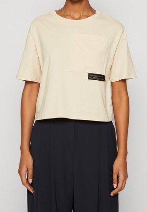 Person trägt ein beigefarbenes, kurz geschnittenes T-Shirt mit Brusttasche und schwarzem Etikett, kombiniert mit hoch taillierten schwarzen weiten Hosen.