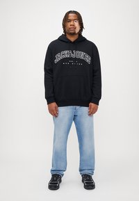 Sort hættejakke med hvid "JACK&JONES" logo, frontlomme, parret med lyseblå jeans og sorte sportssko. Model står med fronten vendt fremad.