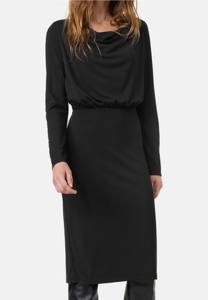 Frau trägt ein schwarzes Midi-Kleid mit langen Ärmeln, einem drapierten Ausschnitt und taillierter Passform, steht vor einem schlichten Hintergrund.