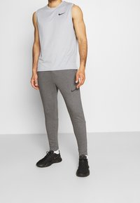 Graues ärmelloses Nike-T-Shirt kombiniert mit grauen schmalen Jogginghosen; beide aus weichem Material. Schwarzes Nike-Logo auf dem T-Shirt und den Jogginghosen. Schwarze Turnschuhe.
