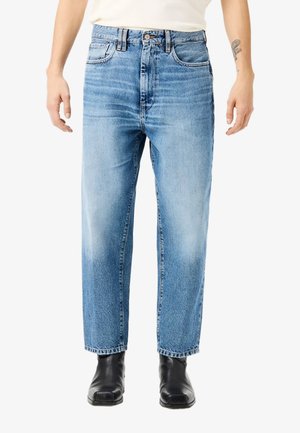 Persona che indossa jeans azzurri a gamba dritta, stivali neri con punta quadrata e una maglietta bianca infilata, in piedi davanti a uno sfondo bianco.