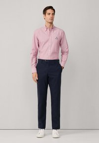 Hombre con una camisa de botones rosa claro con un emblema bordado, pantalones azul marino y zapatillas blancas, de pie contra un fondo liso.
