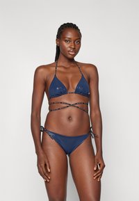 Set bikini blu navy con top triangolare e spalline incrociate e slip con lacci laterali. Texture liscia con dettagli del logo.